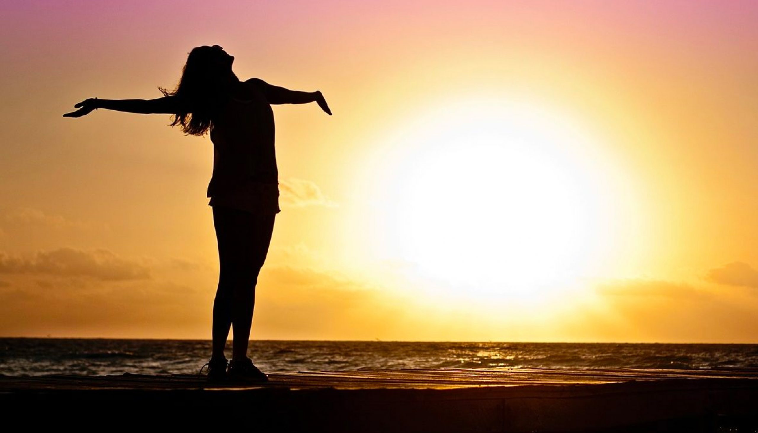 太陽をバックに海辺で両手を広げる女性のシルエット
